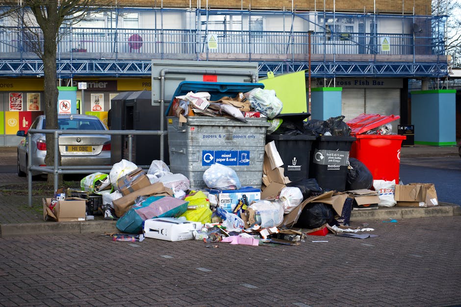 A middle-aged man with dark hair, wearing a black T-shirt with white text and black pants, is seen emptying a white plastic bag filled with rubbish into a silver, stainless steel recycling bin situate